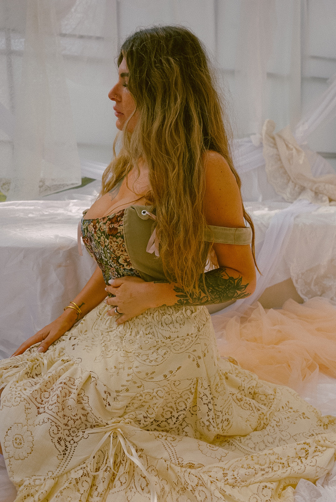 Woman in a lace dress sitting in a softly lit room with white curtains.