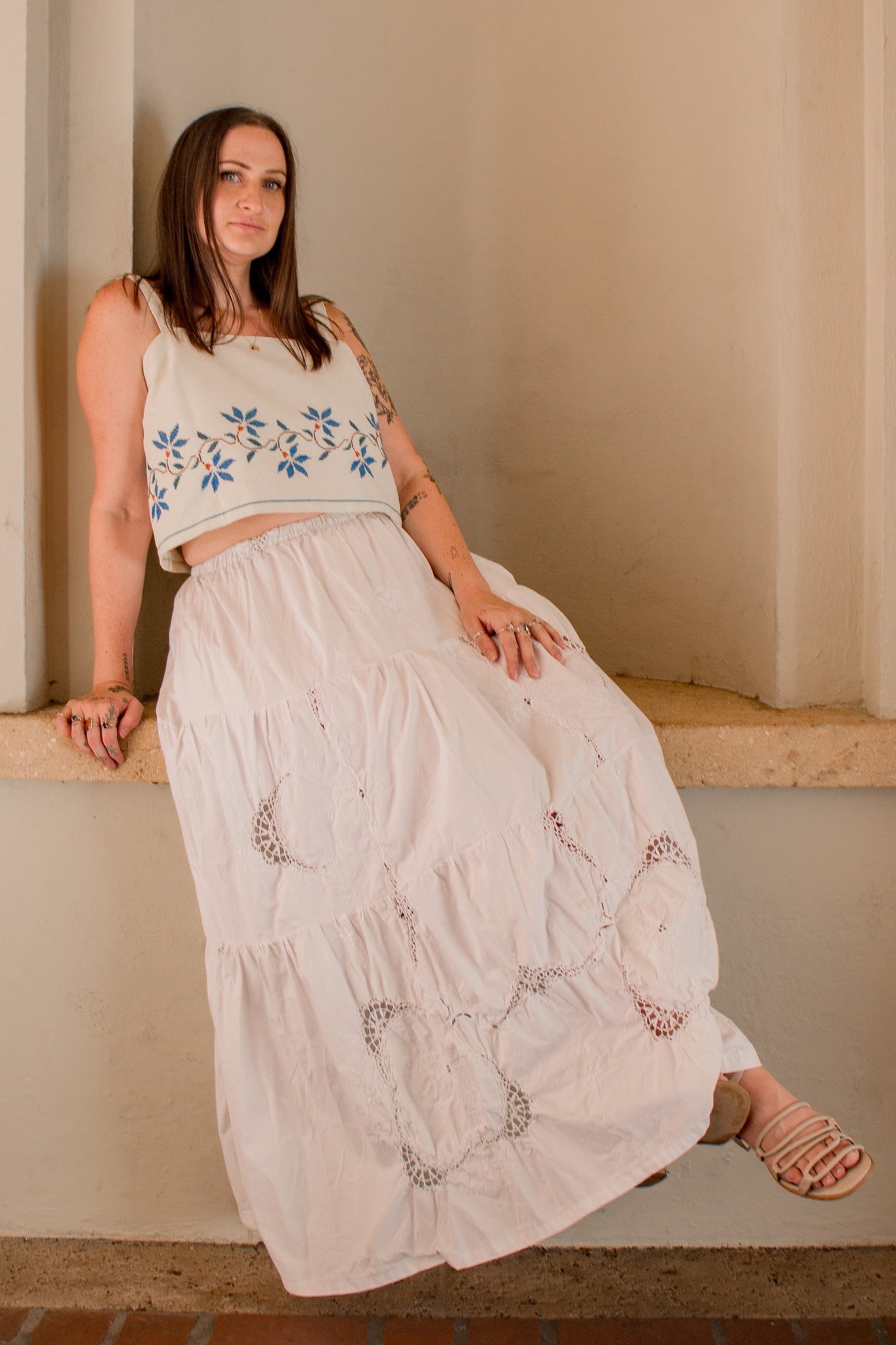 Woman in a white embroidered dress sitting on a ledge against a beige wall.
